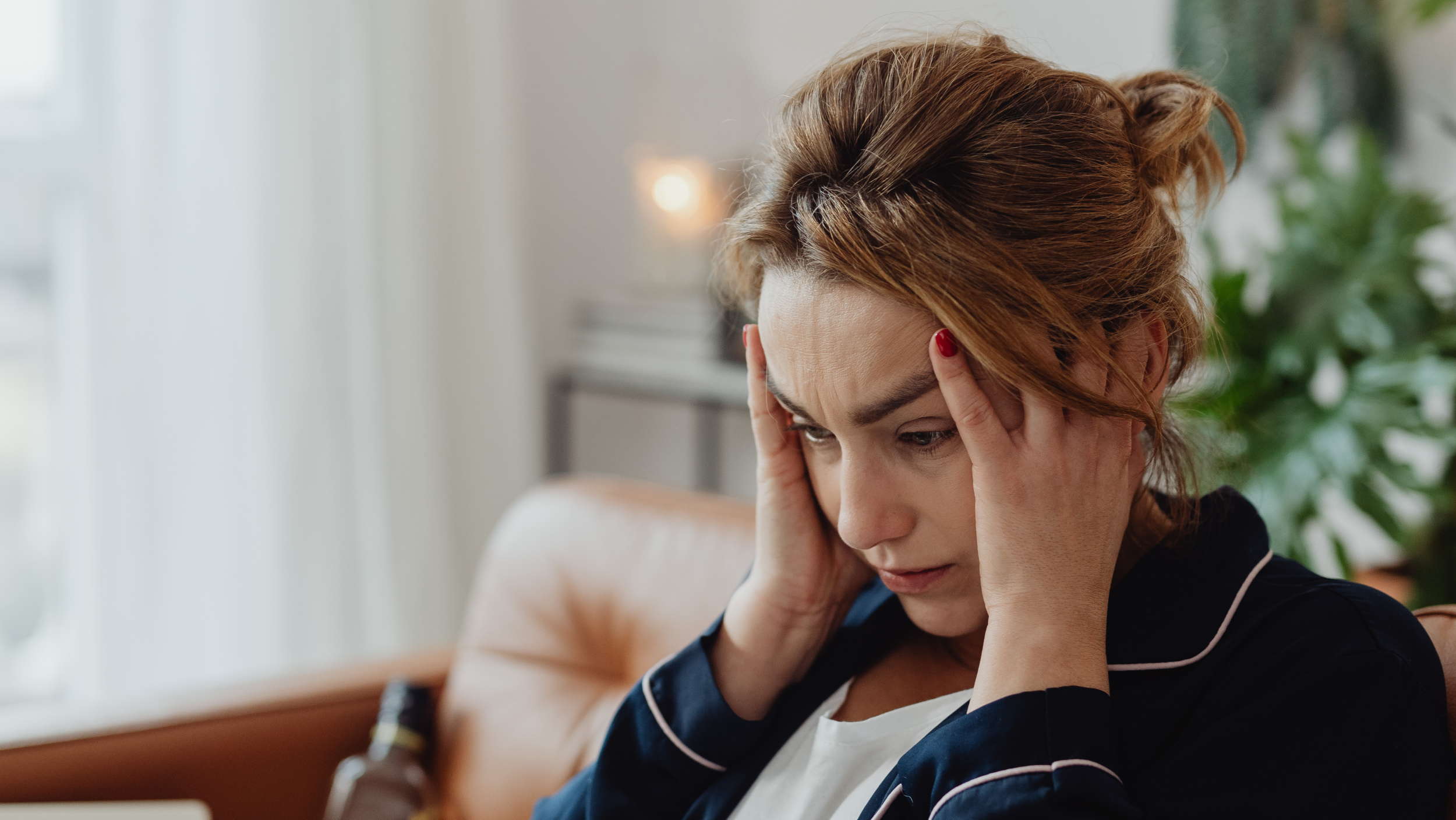Eine gestresste Frau sitzt im Wohnzimmer und hält ihre Hände an die Schläfe / Kopf.